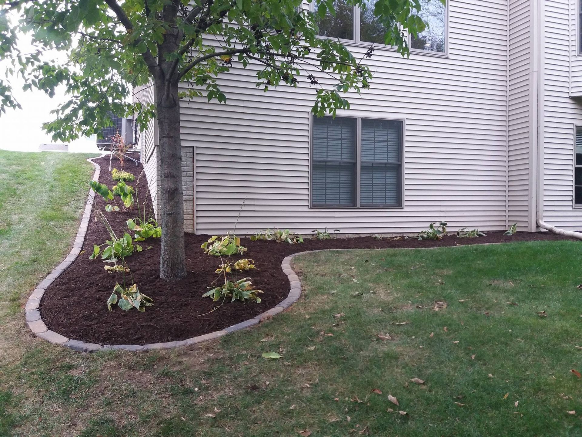 House exterior with a mulched flower bed, tree, and curved stone border beside a lawn