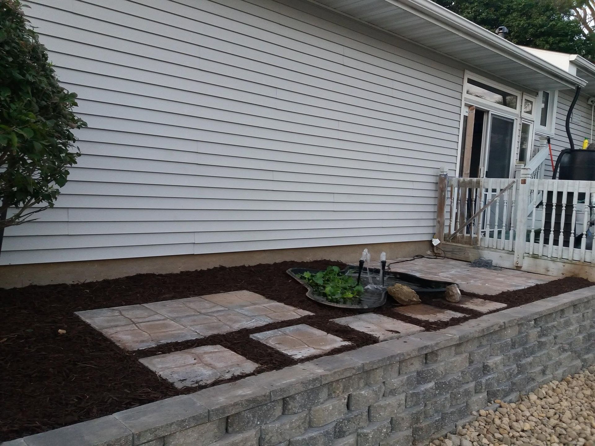 Side yard with stepping stones, mulch beds, and a stone retaining wall beside a white house and porch.