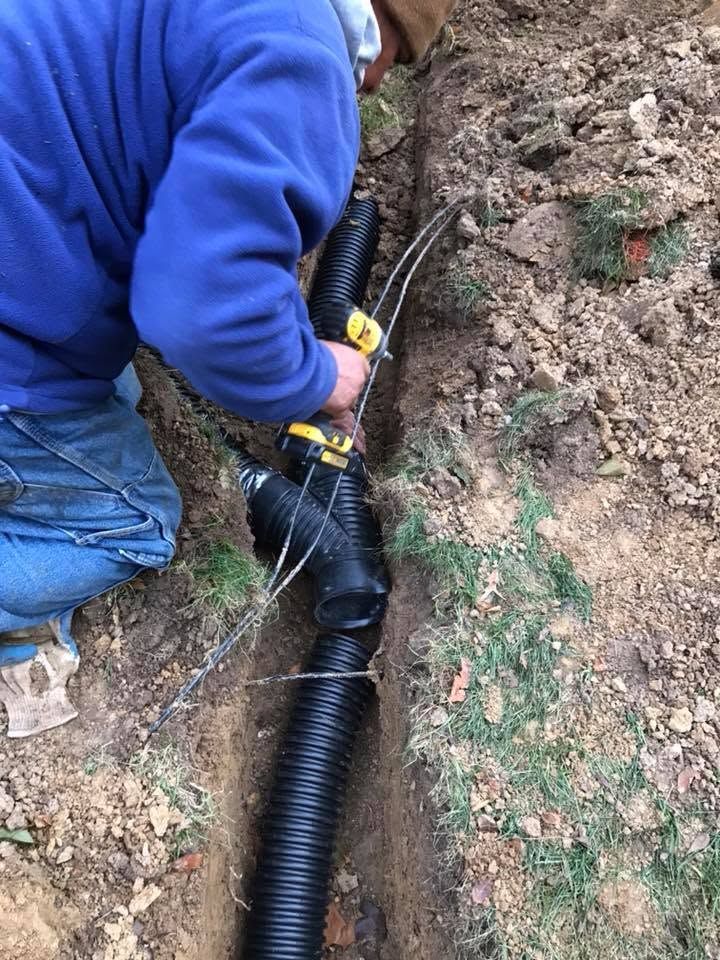 Worker in blue hoodie installing black conduit in a muddy trench near exposed soil and rocks