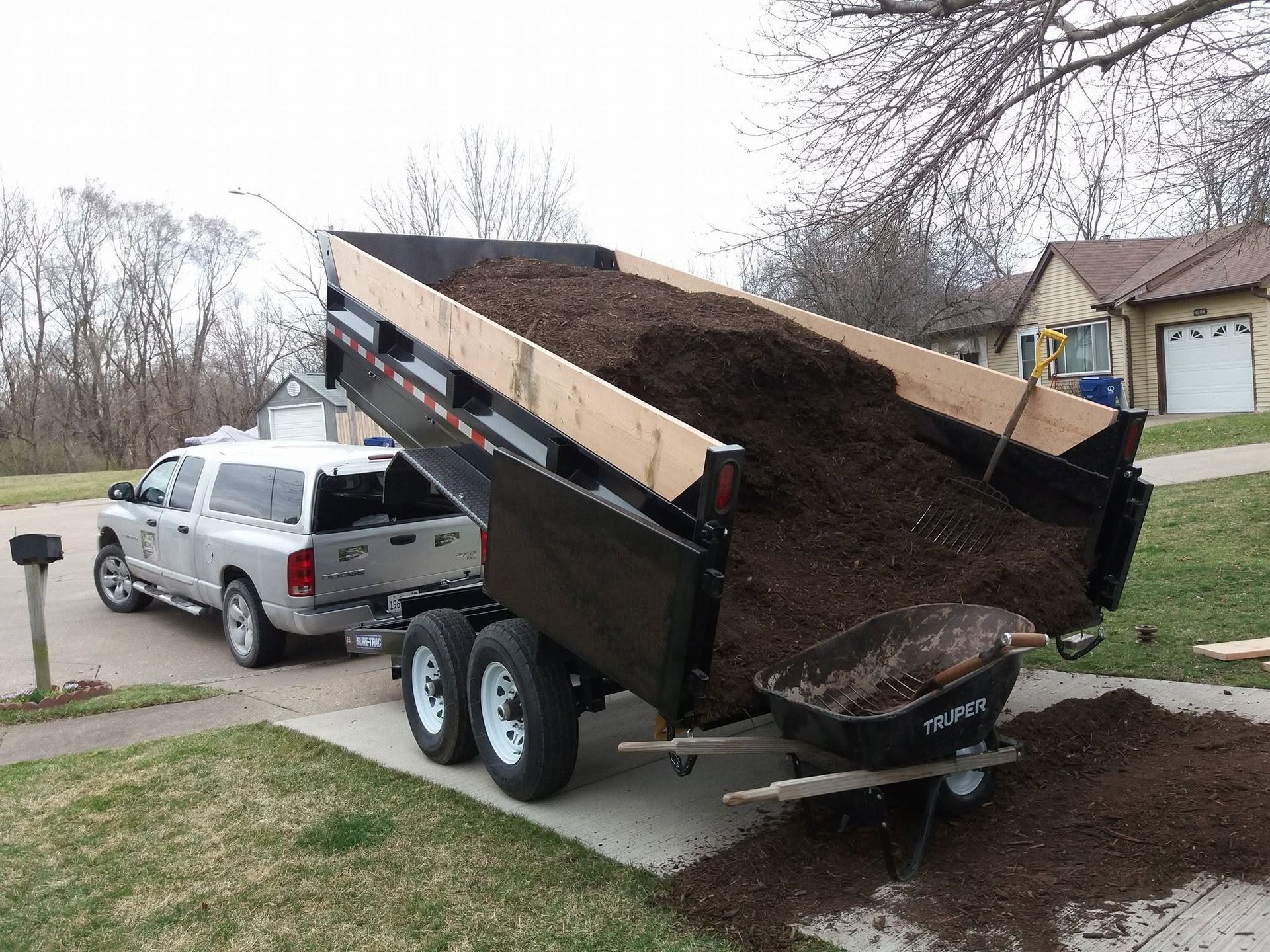 Pickup truck dumping a load of dark soil from a black trailer onto a driveway