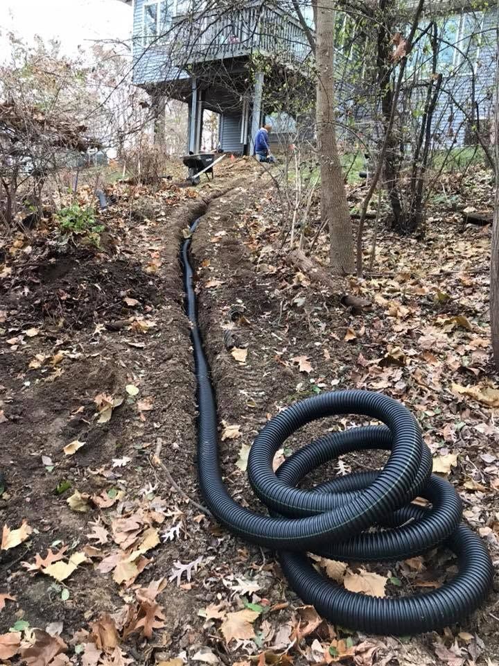 Black hose runs uphill through leaf-covered yard to a porch, with two people near the house.