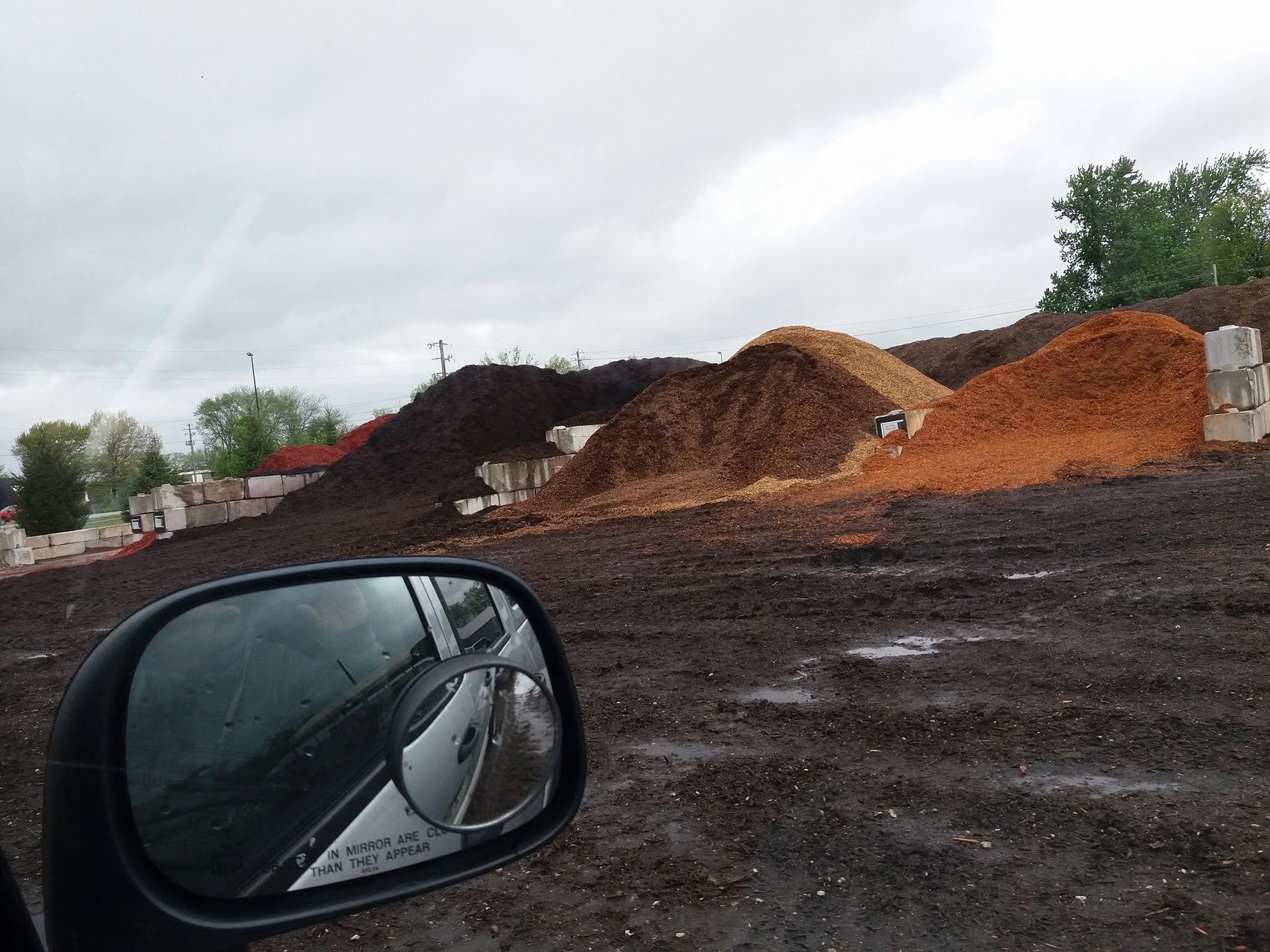 Car side mirror overlooking piles of dark soil and orange gravel at a construction site