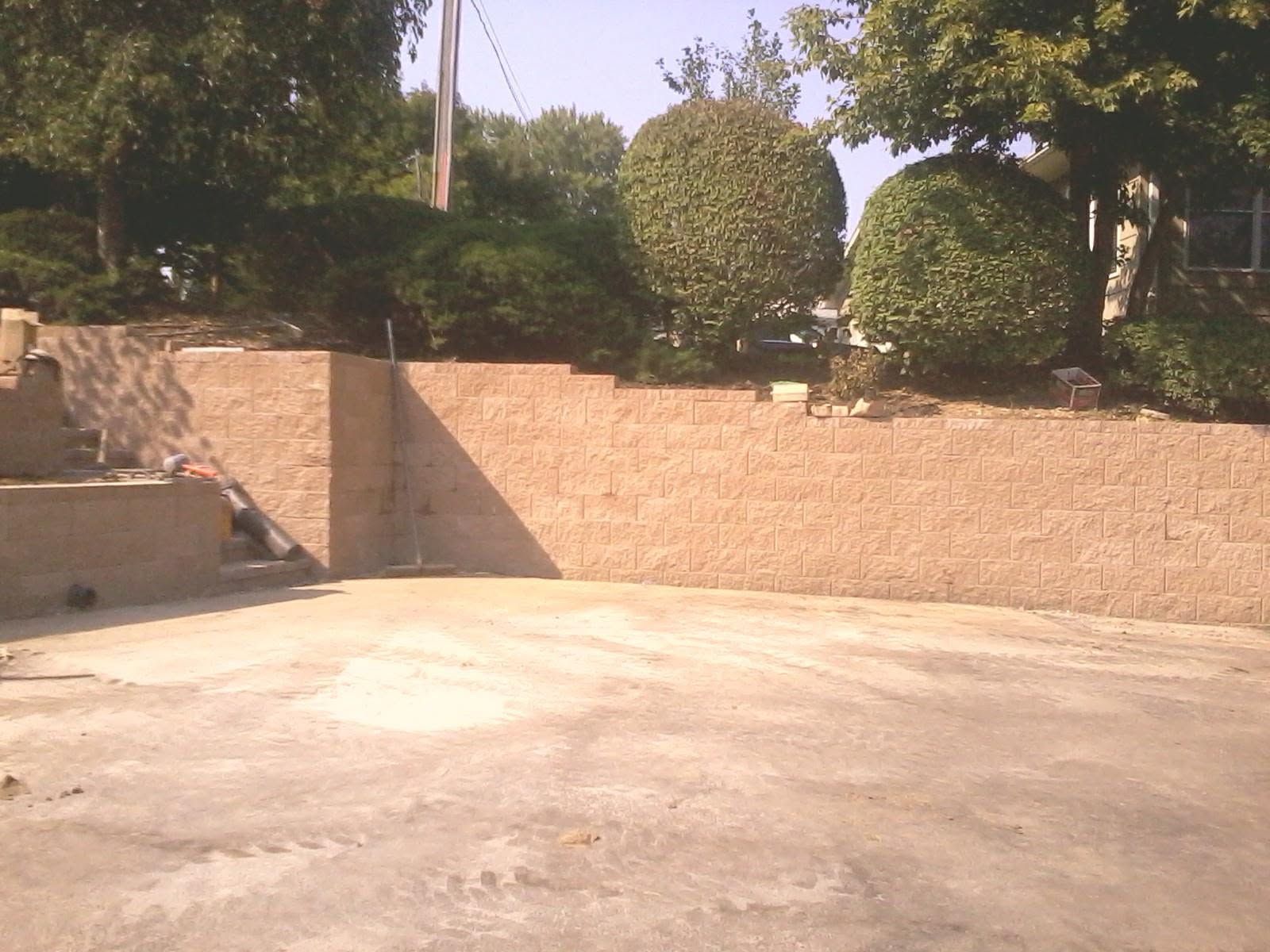 Empty concrete courtyard beside a tan brick wall with trimmed bushes and trees in bright sunlight