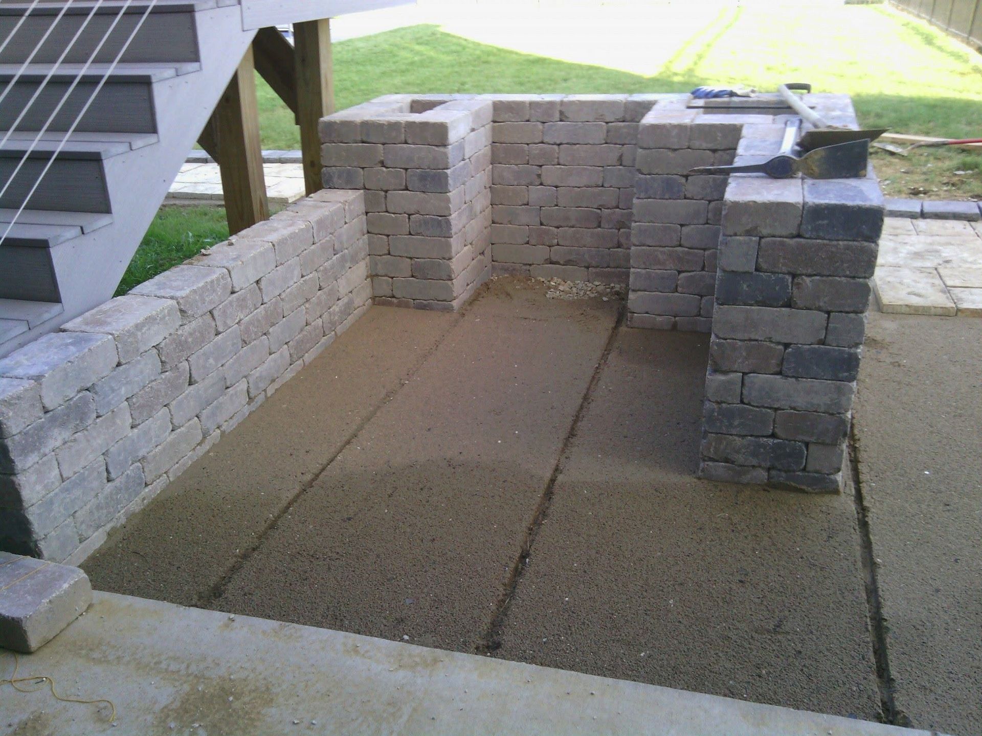 Small brick-and-cinder-block patio enclosure under outdoor stairs beside a grassy yard.