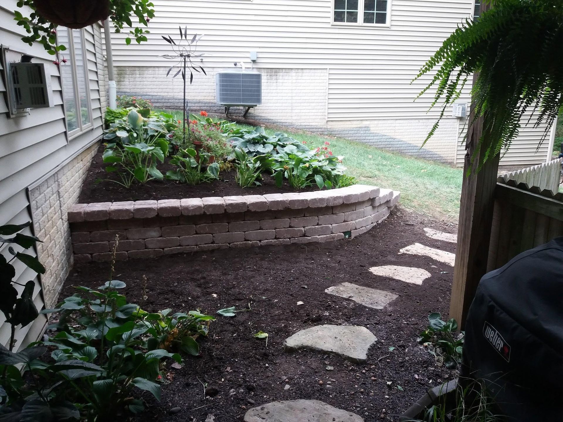Backyard garden bed beside house with stone edging, stepping stones, and green plants