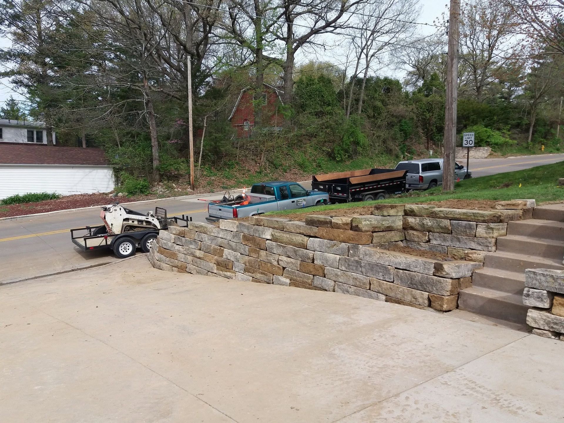 Stone retaining wall under construction beside a driveway, with work trailer and equipment nearby.