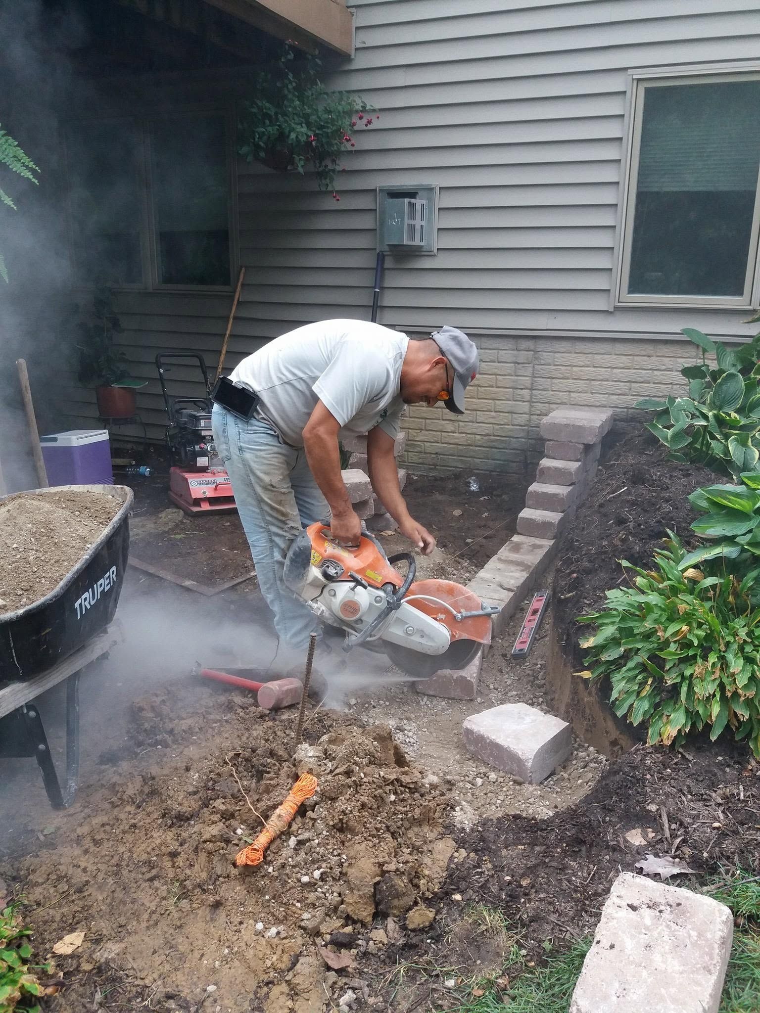 Person using a chainsaw to cut a concrete slab beside a house, with dust and debris around them