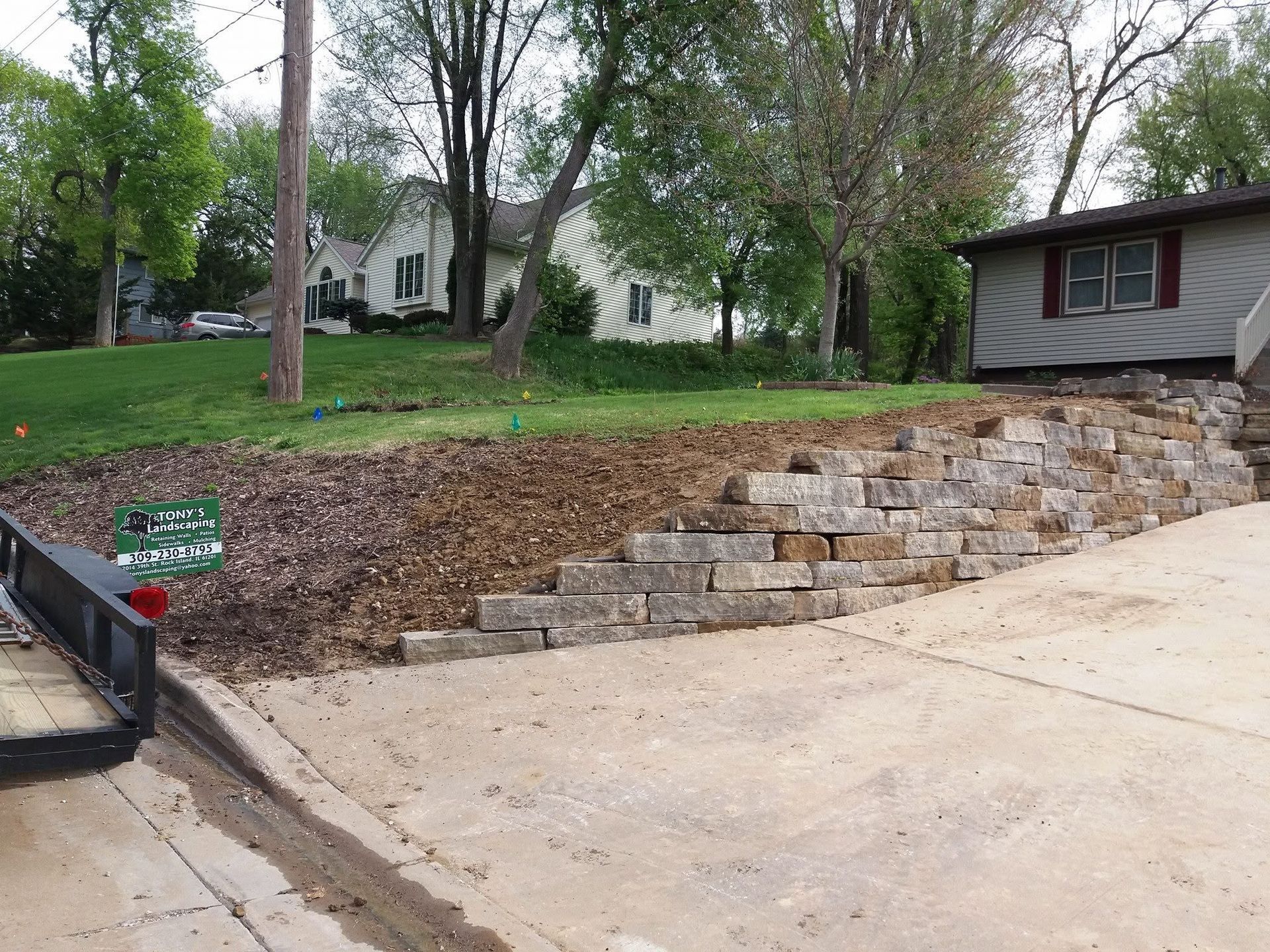 Retaining wall with stone steps beside a driveway, leading to a grassy yard and house