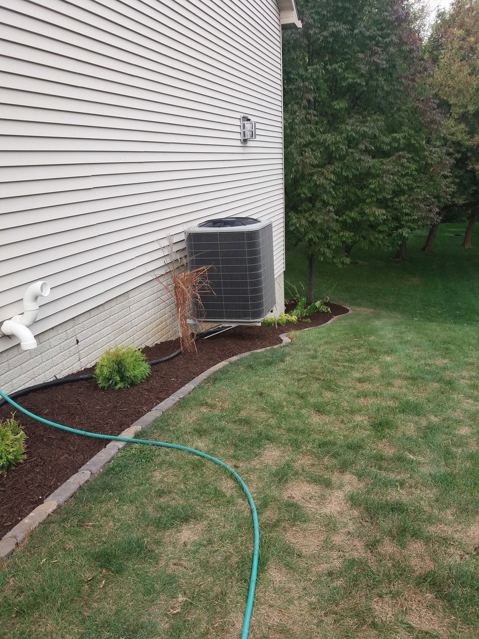 House side with air conditioner unit, garden hose, mulch bed, and lawn next to a tall hedge.