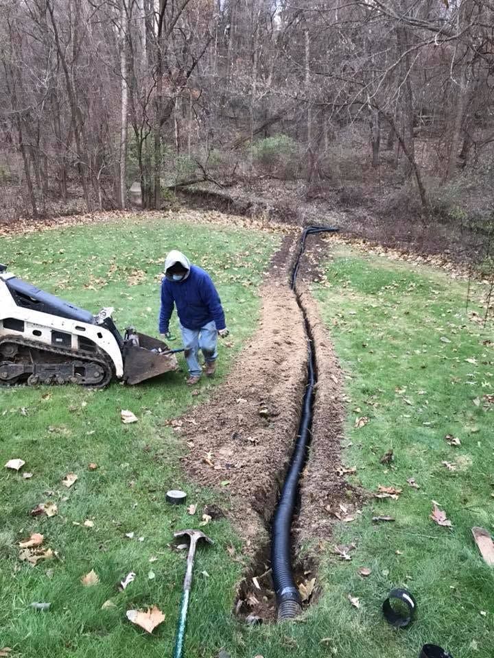 Worker beside skid steer installing black drainage pipe in a muddy yard.