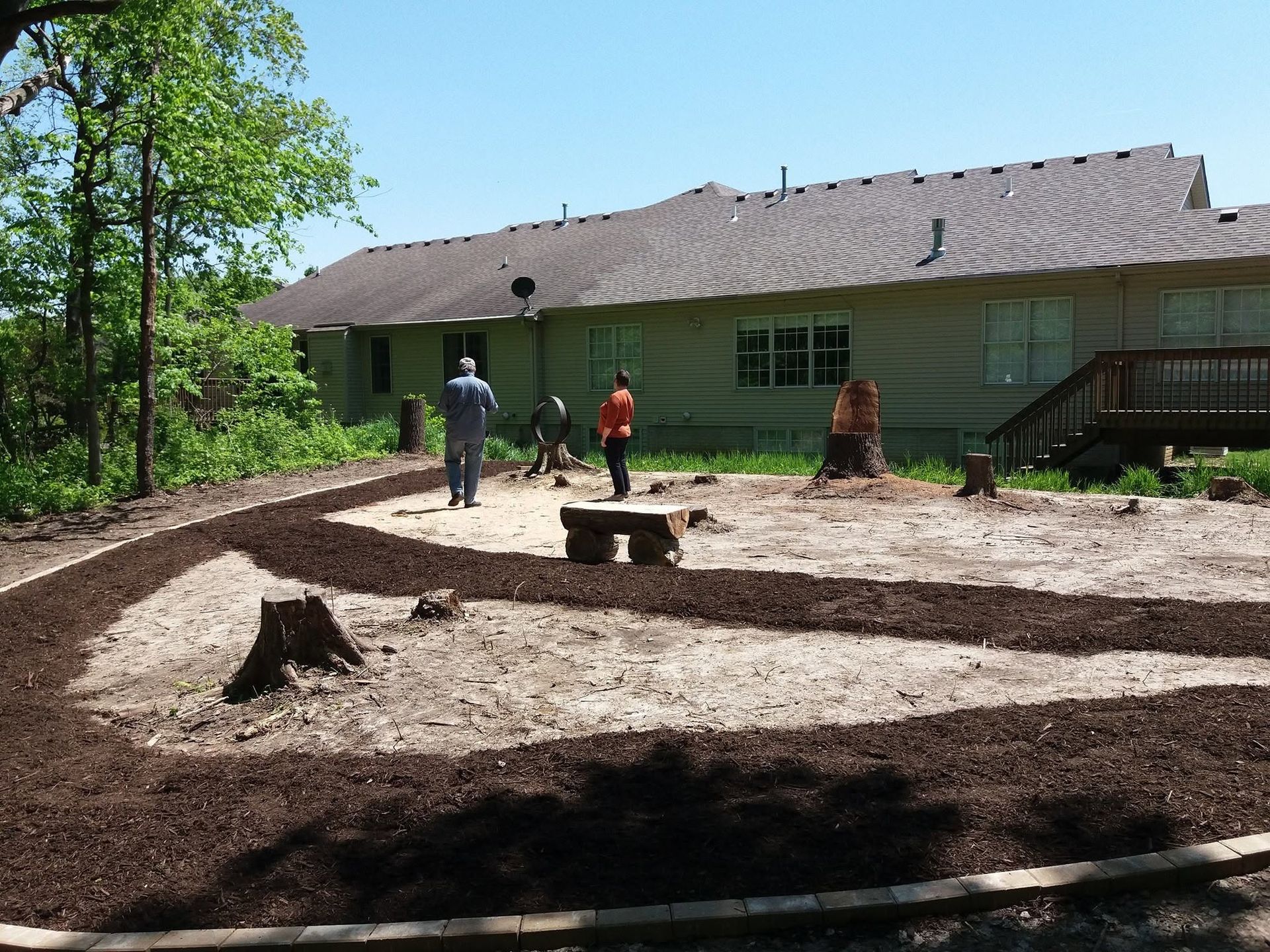 Workers landscaping a yard beside a green house, with fresh mulch and a small fire pit area