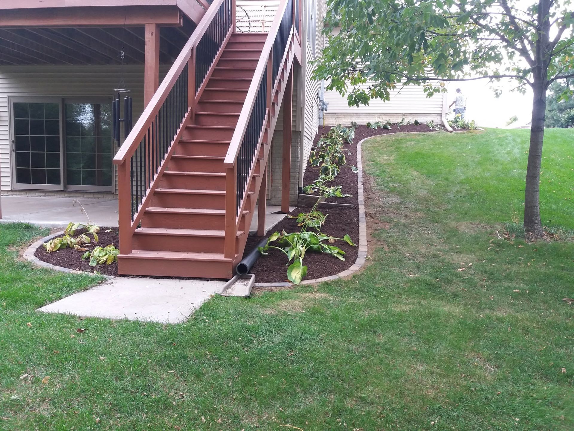 Brown wooden outdoor stairs beside a house, with a concrete path and green lawn.