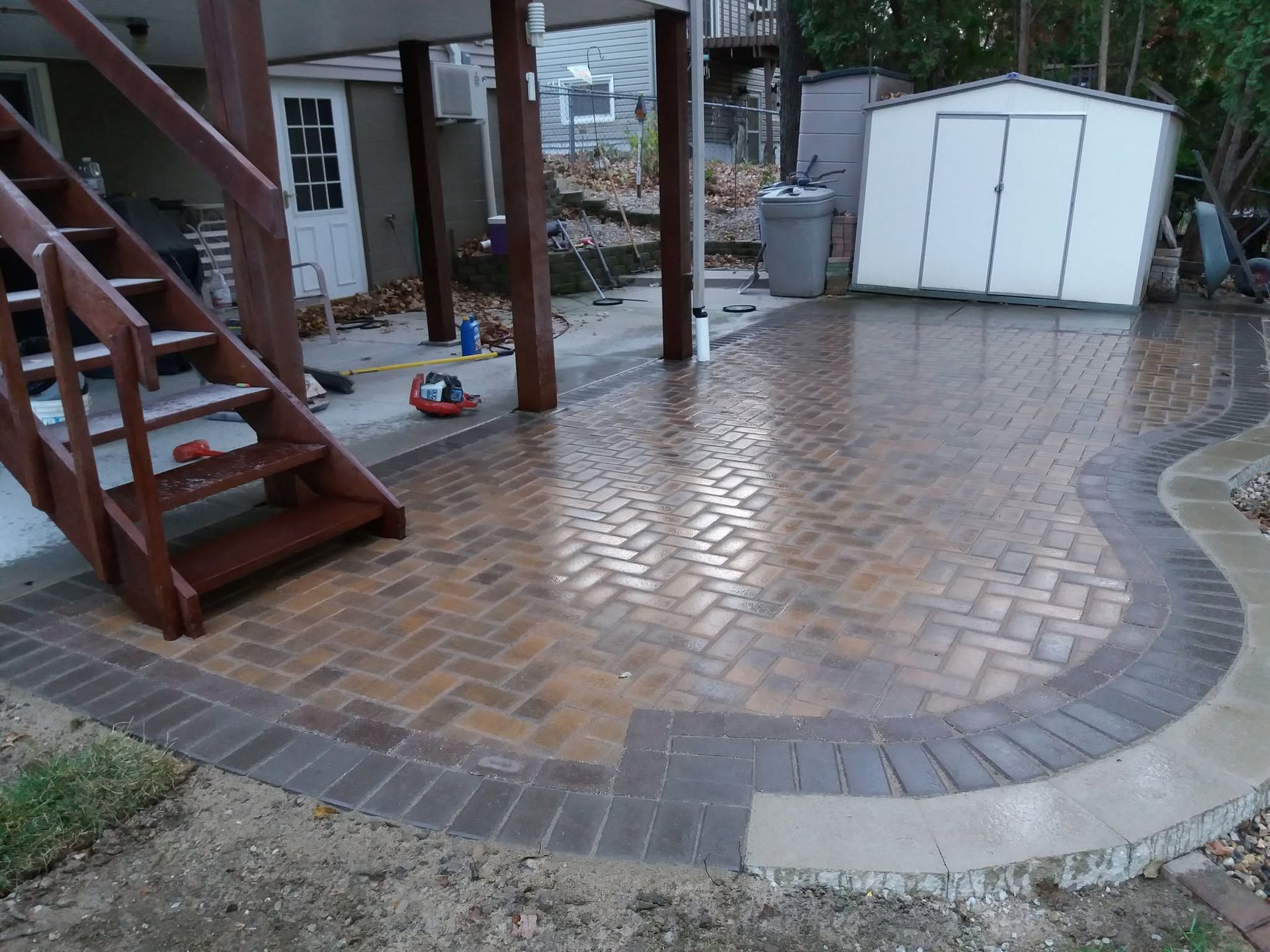 Brick patio under a wooden deck beside a white shed, with curved stone edging.