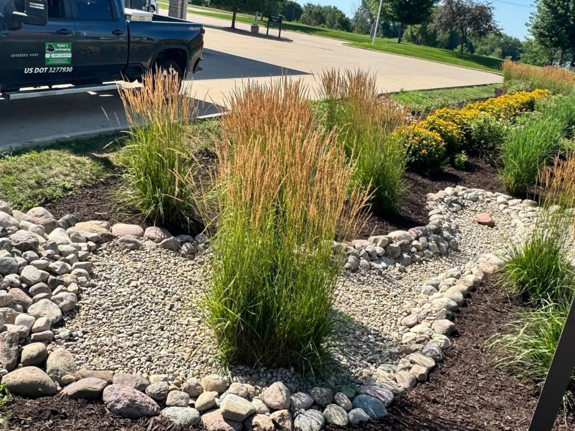 Mulched landscaping bed with rocks and ornamental grasses beside a driveway and parked vehicle