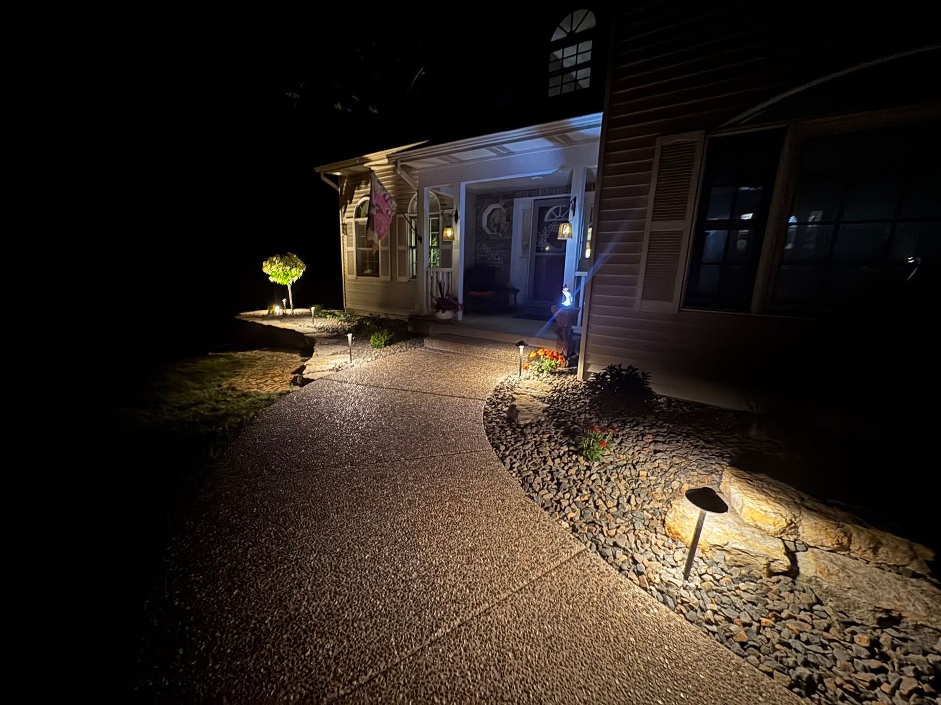 Dimly lit gravel walkway leading to a house entrance at night, with pathway lights and a porch lamp.