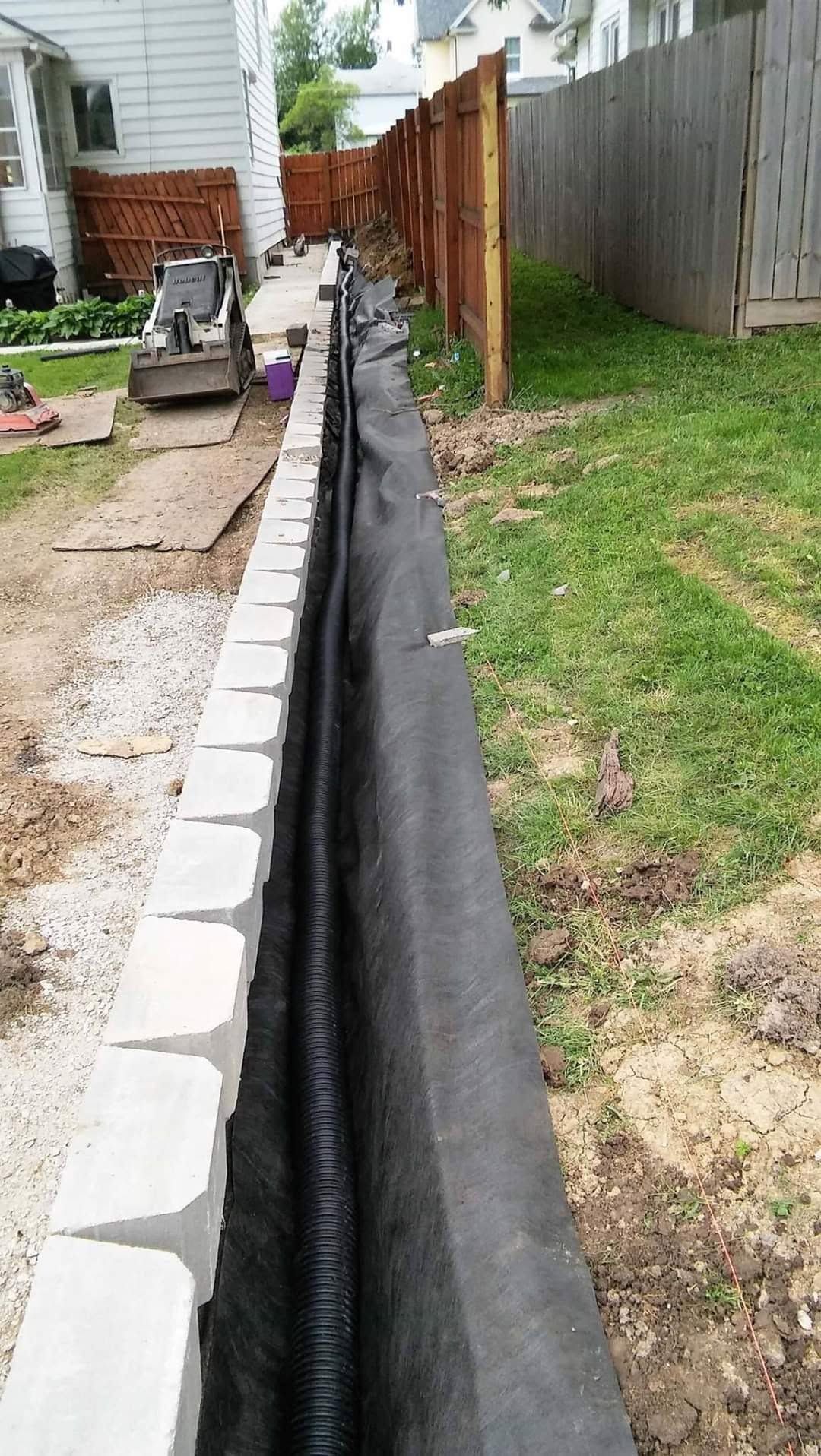 Construction site with retaining wall blocks and black fabric lining a narrow trench beside a house and fence