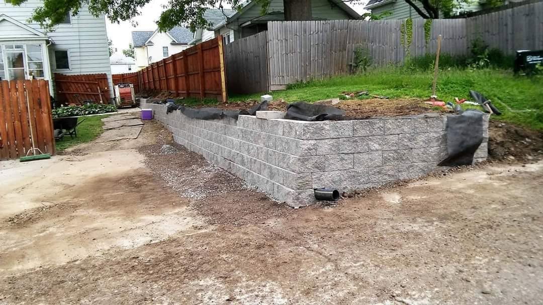 Backyard with a newly built gray stone retaining wall and dirt driveway beside a wooden fence