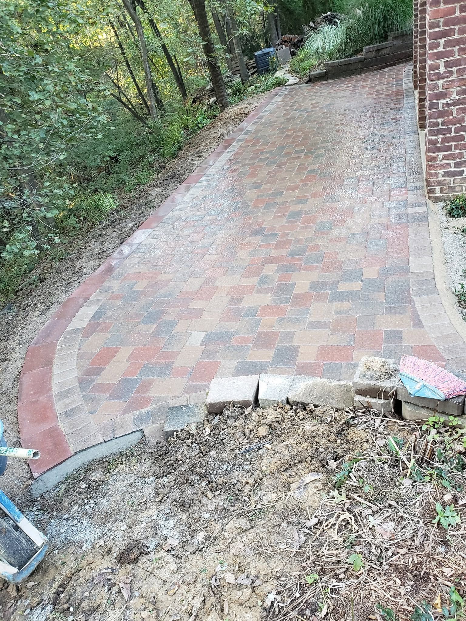 Brick-paved walkway beside a house, bordered by trees and a curved concrete edge with gravel foreground