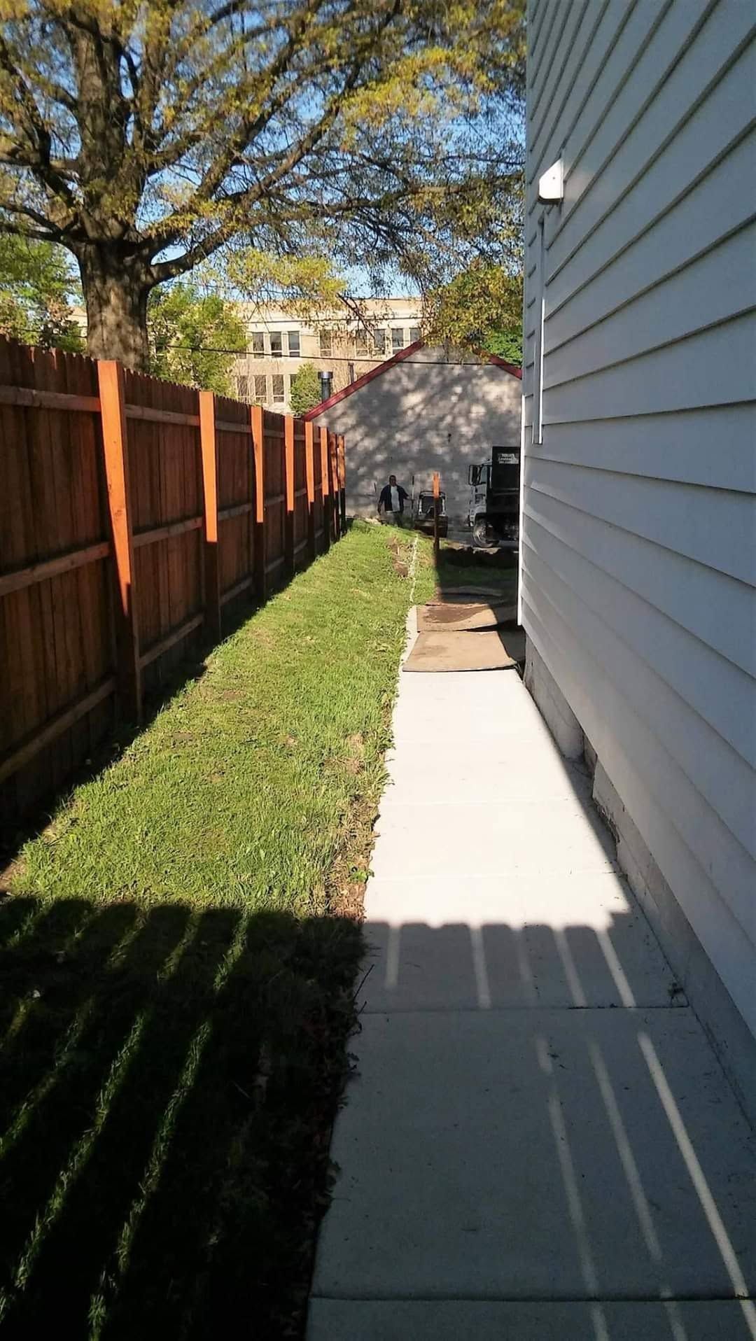 Narrow concrete walkway beside a house, bordered by grass and a wooden fence in bright sunlight.