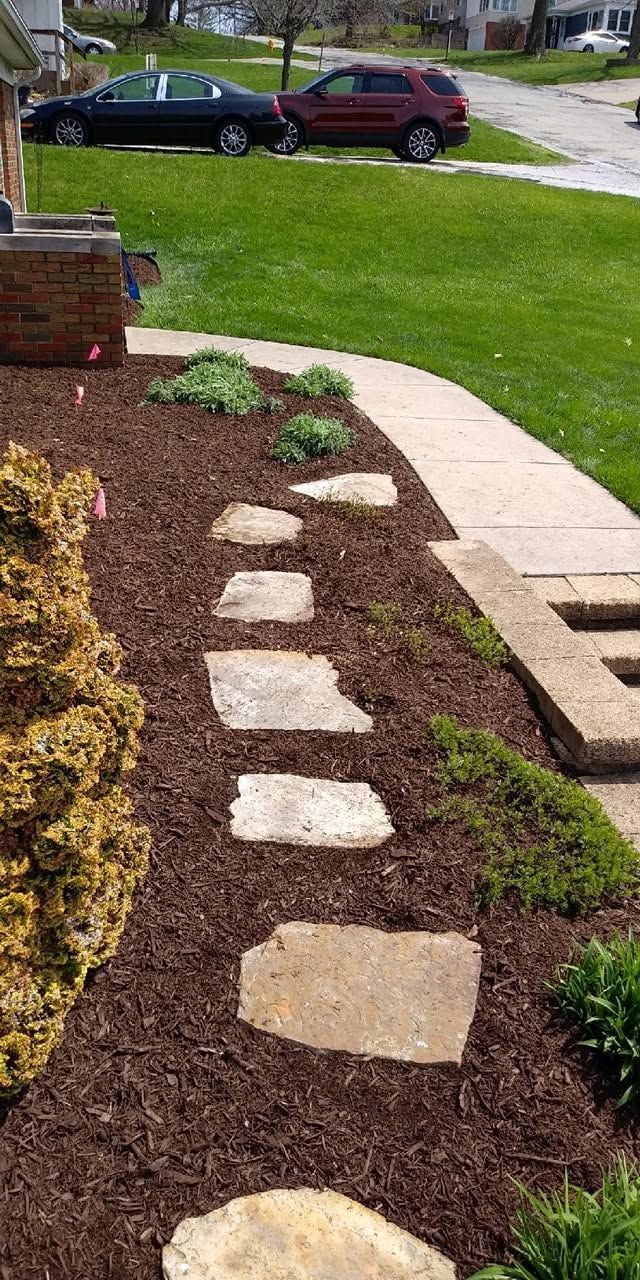 Stepping-stone path through mulch in a front yard toward a sidewalk and parked cars