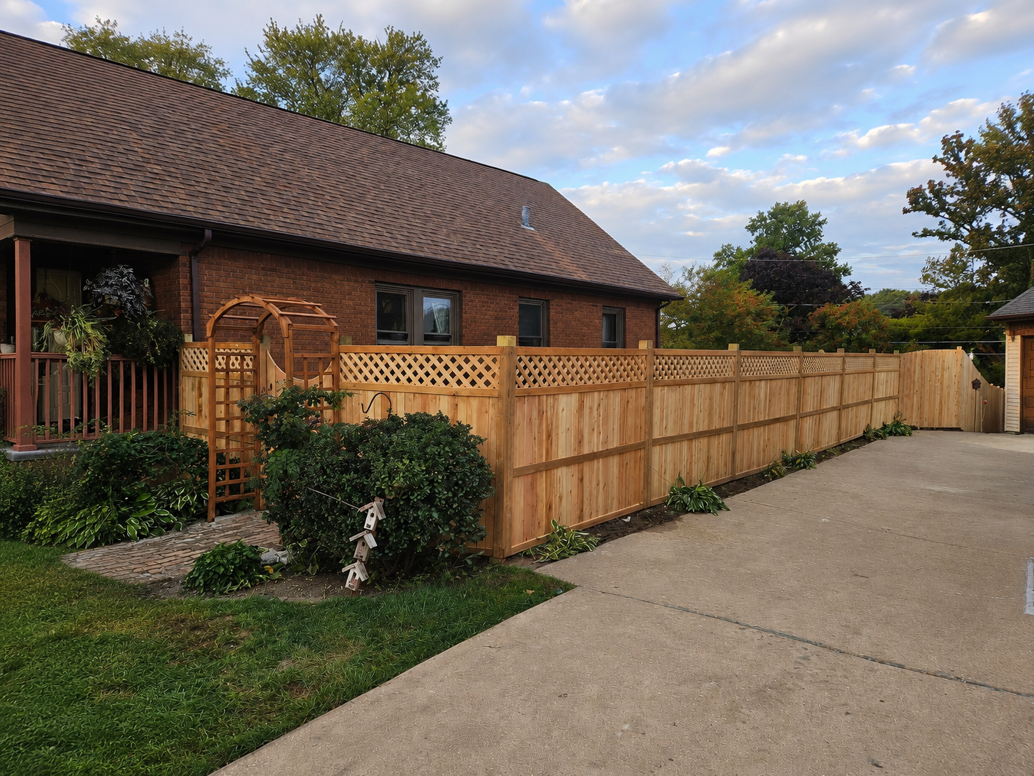 Tilted view of a stone sidewalk beside a wooden fence, shrubs, and a building with a tiled roof under a cloudy sky
