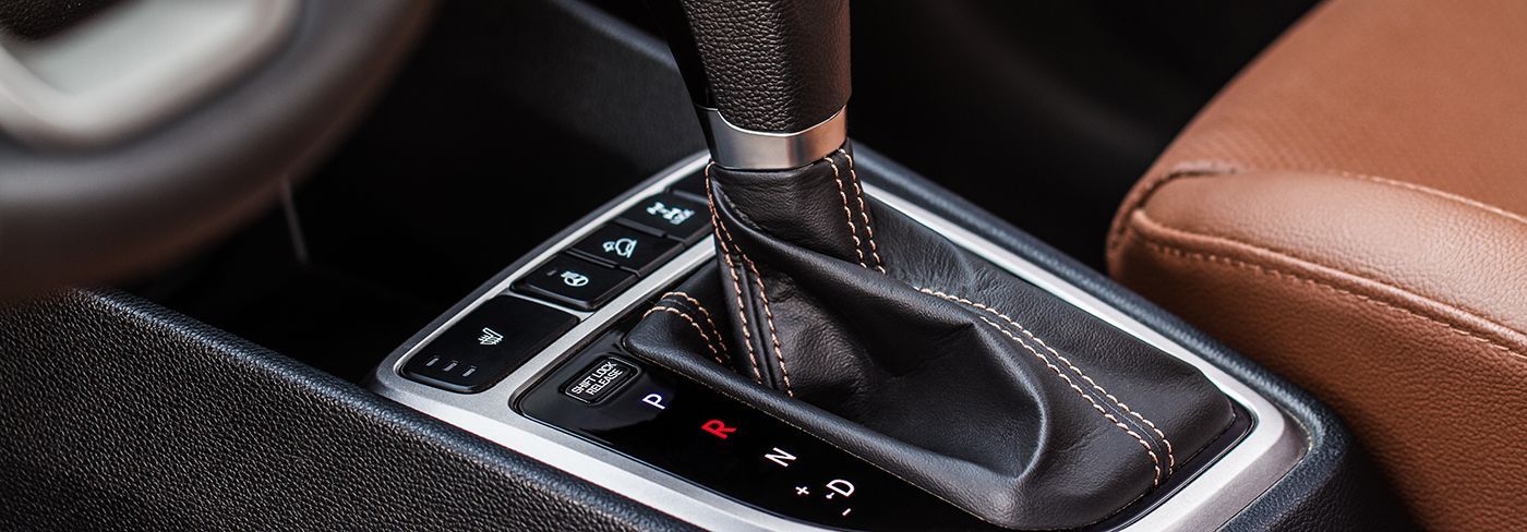 Close-up of a car's gear shift lever, in the center console. Brown leather seat visible in the background.