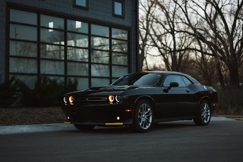 A black Dodge Challenger parked on a paved driveway in front of a modern garage with large glass panels at dusk.