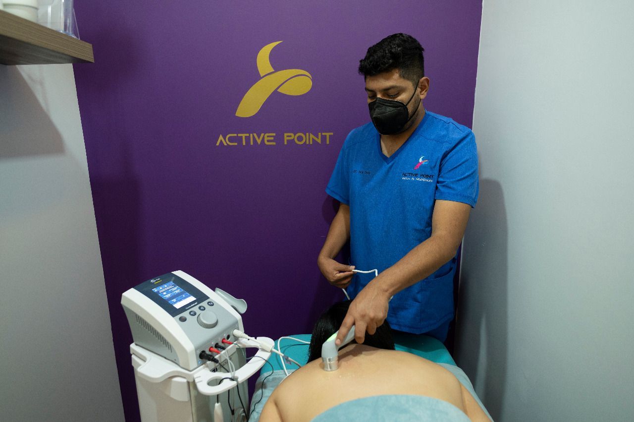 Un hombre con uniforme azul usa un ecógrafo en la espalda de un paciente en una consulta de fisioterapia. Pared morada con logotipo.