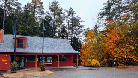 Red cabin-style building with a gray roof beside bright autumn trees and a gravel lot