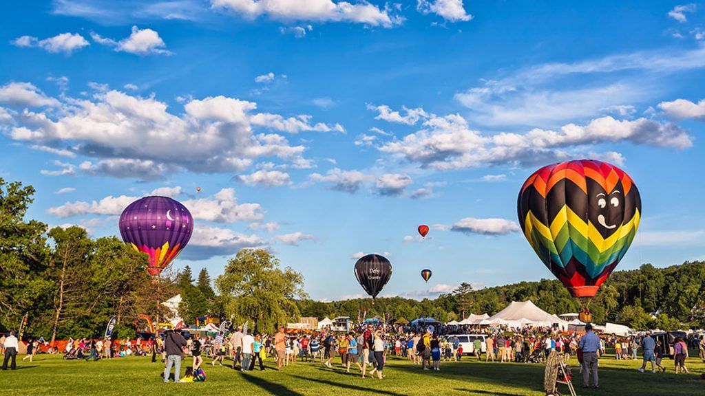 Crowd in a park watching colorful hot air balloons launch under a blue sky