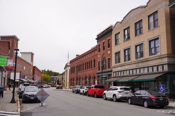 Small-town street with brick storefronts and parked cars on a cloudy day