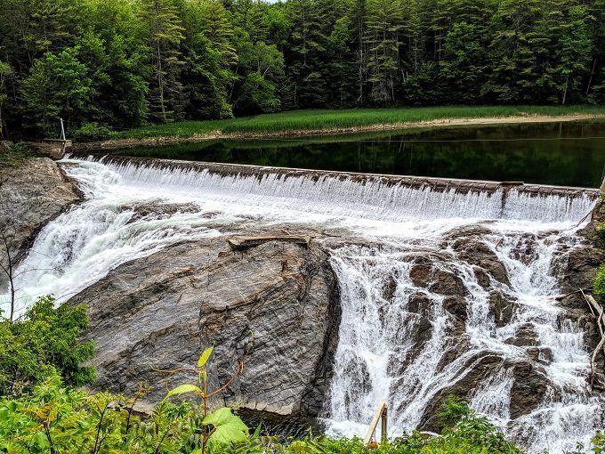 Water cascading over a low dam into a rocky stream, surrounded by lush green forest.