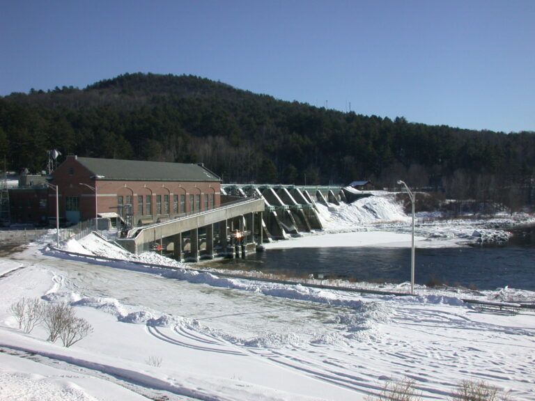 Snow-covered dam and power plant beside a river, with a forested hill in the background.