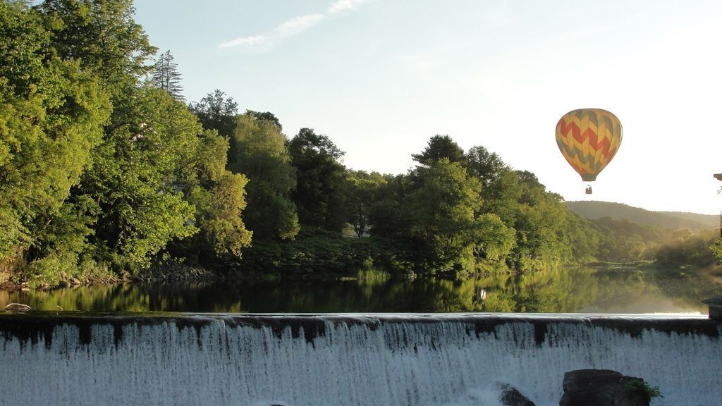 Tree-lined river with a low waterfall at sunset, with a colorful hot air balloon in the sky