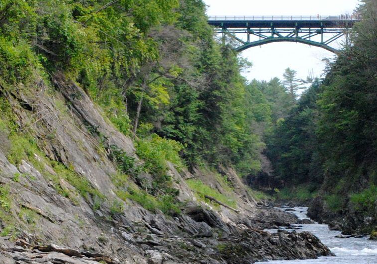 Rocky river gorge beneath a steel bridge, with green forested cliffs and rushing water.
