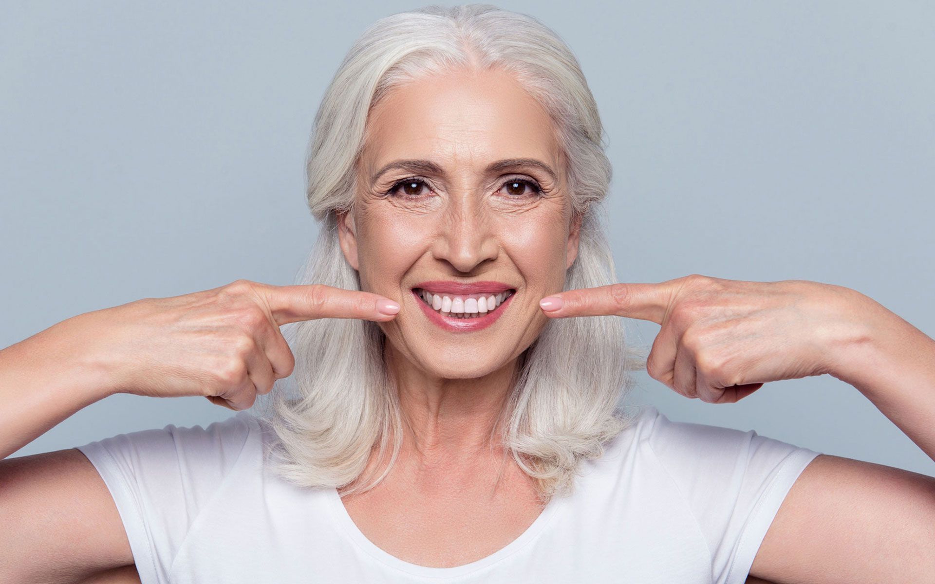 An older woman is smiling and pointing at her teeth.