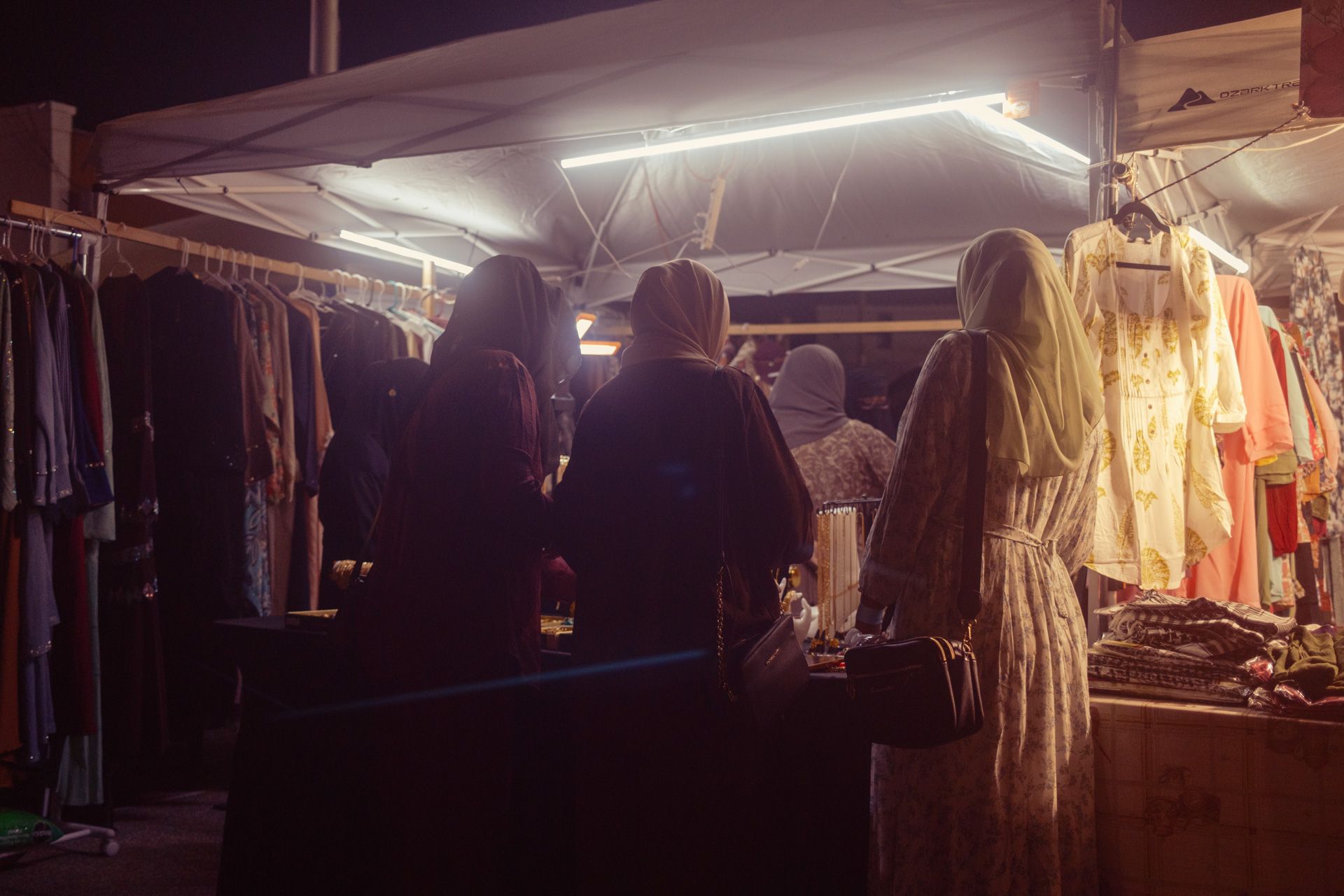 A trio of women with their heads covered gather around a vendor table at a nighttime street market in Detroit, MI.