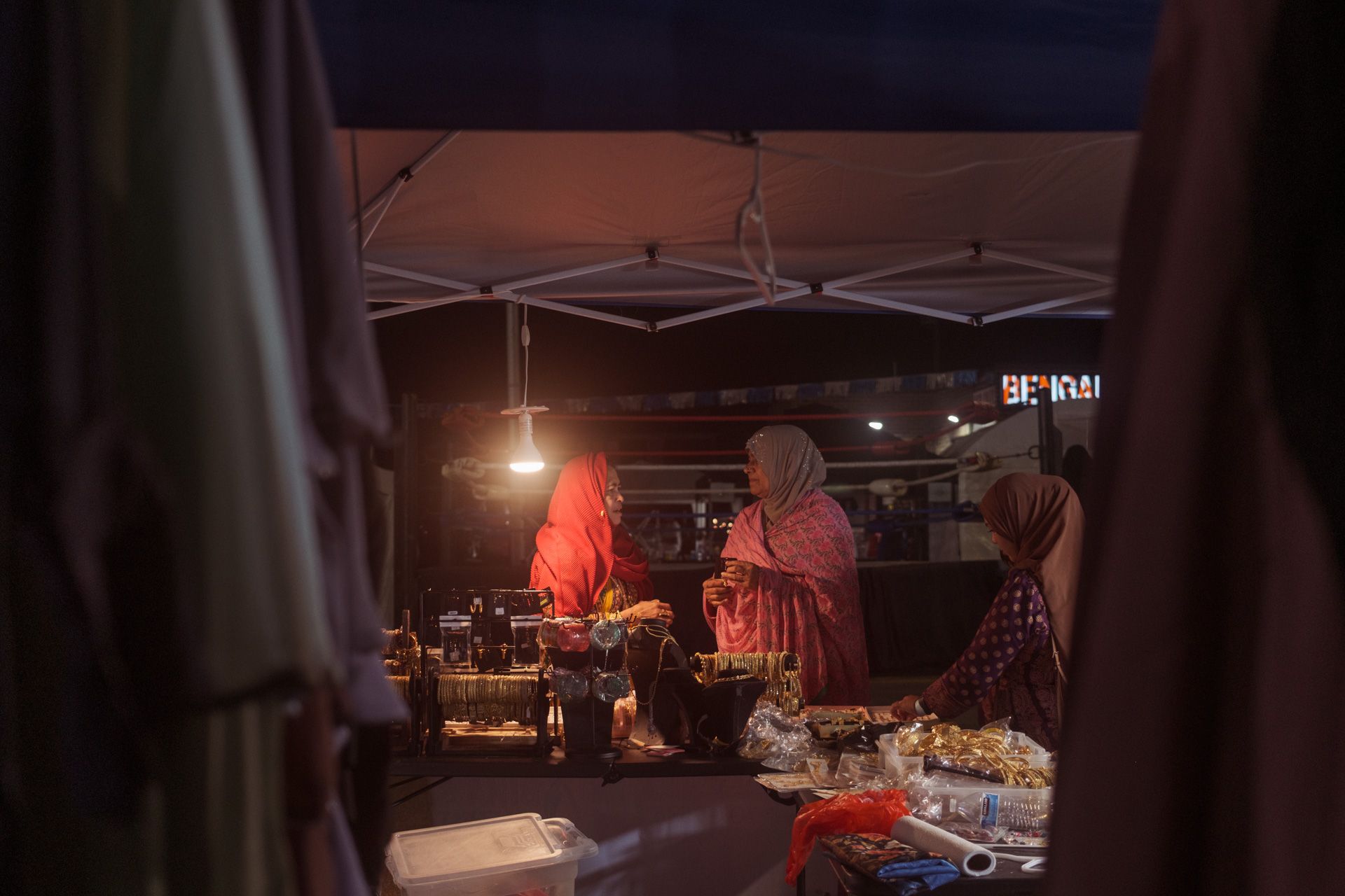 Two women talk under a vendor tent while a younger woman looks on at a night time market. 