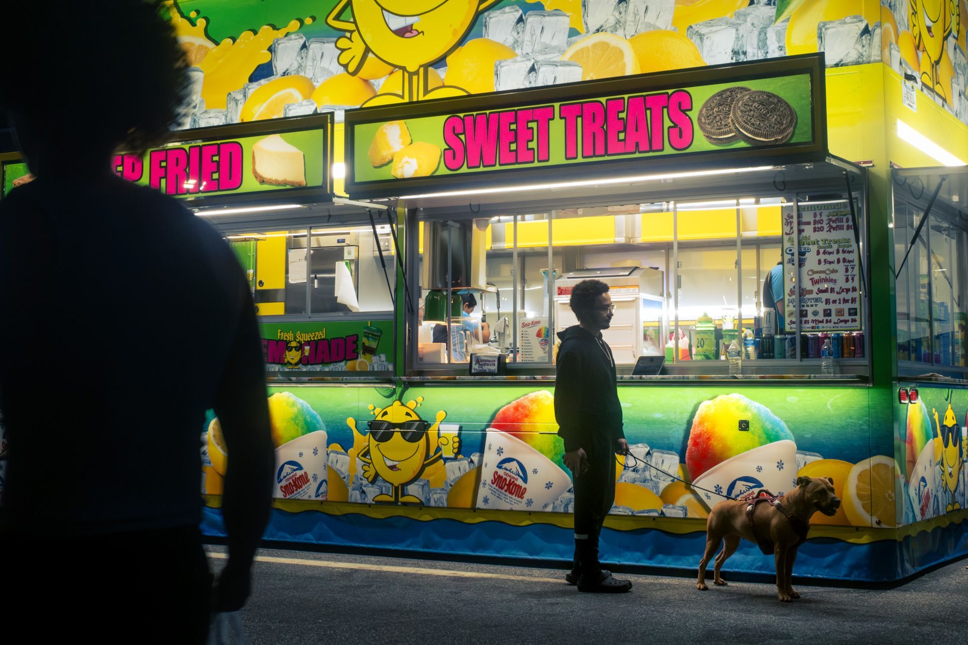 Man and his dog in front of a bright lemonade stand at a night street festival