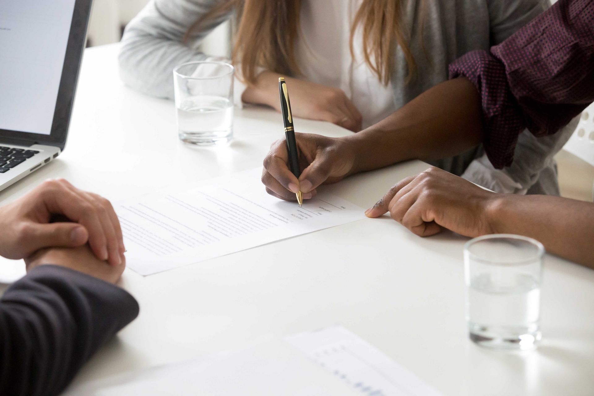 Une personne signe un document à une table, en présence de deux autres personnes. Un ordinateur portable et des verres d'eau se trouvent également sur la table.