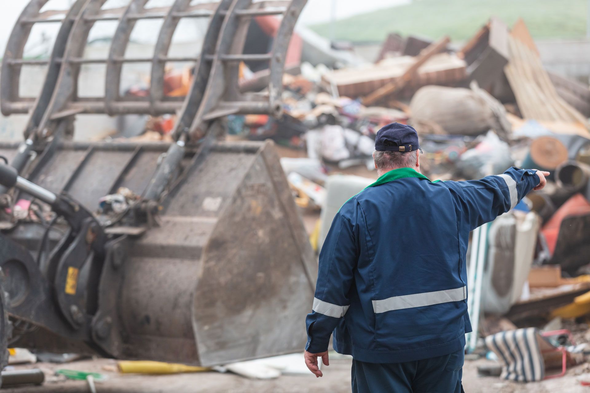 Worker in blue jacket pointing at a collapsed excavator amid rubble and debris.