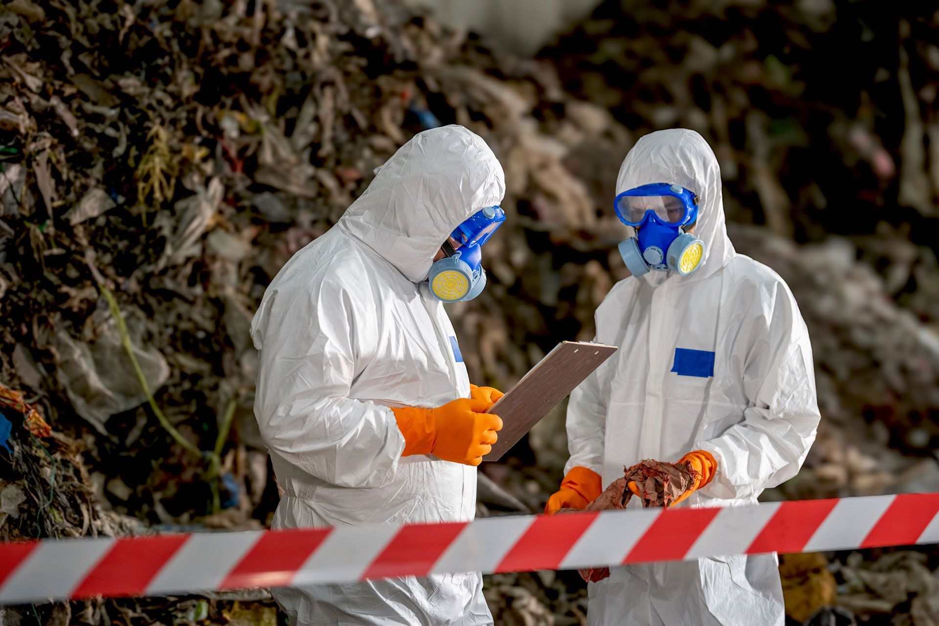 Two hazmat-suited workers inspect a clipboard behind red-and-white tape in a debris-filled area