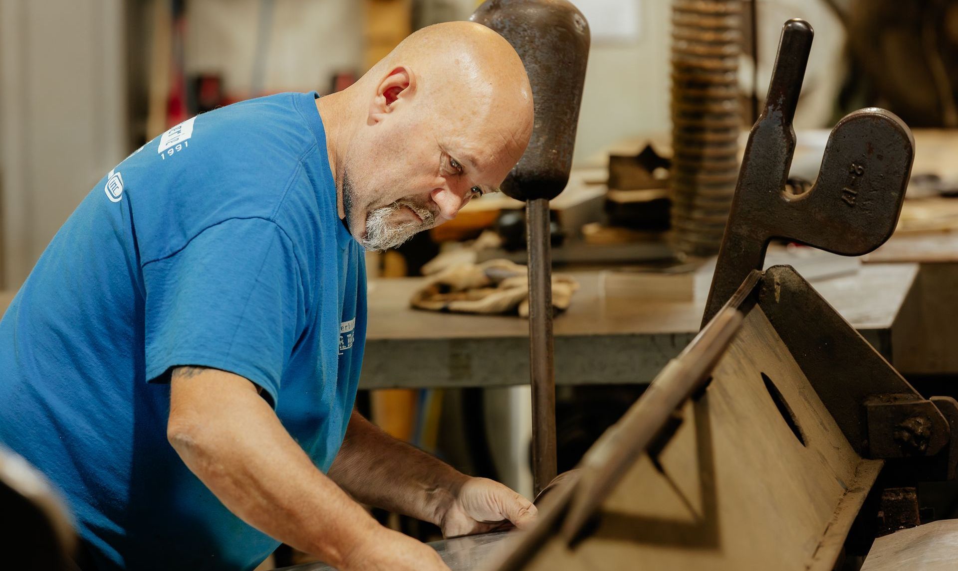Un homme en chemise bleue travaille à un établi, concentré sur sa tâche. Cheveux grisonnants, intérieur.