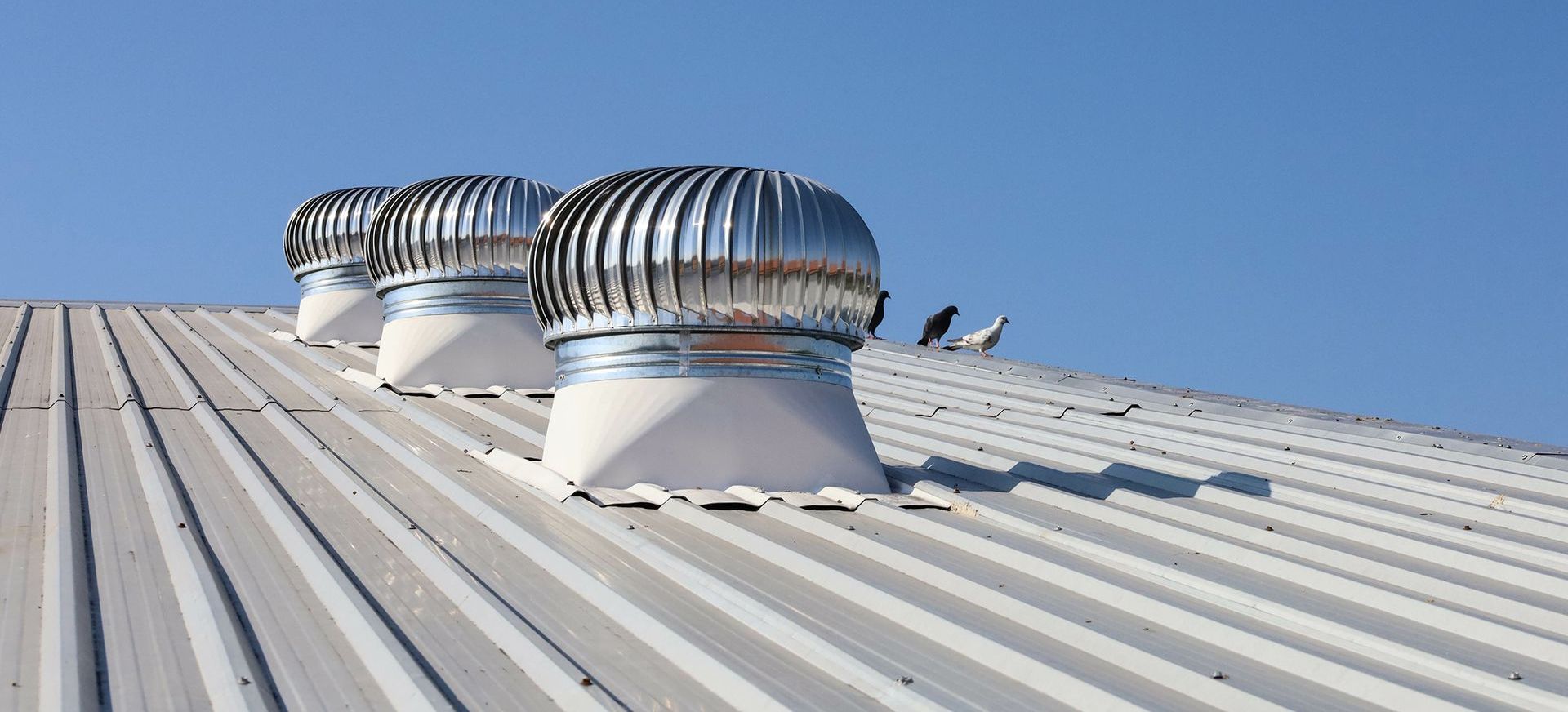 Trois turbines de ventilation sur un toit en tôle ondulée blanche se détachant sur un ciel bleu.