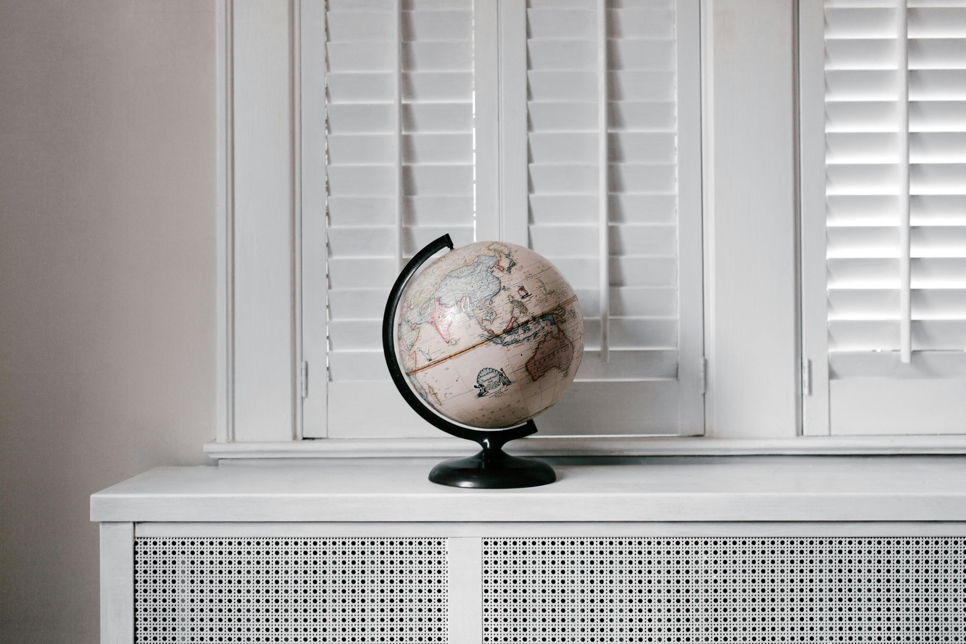Un globe terrestre posé sur un radiateur blanc, devant une fenêtre à volets blancs.