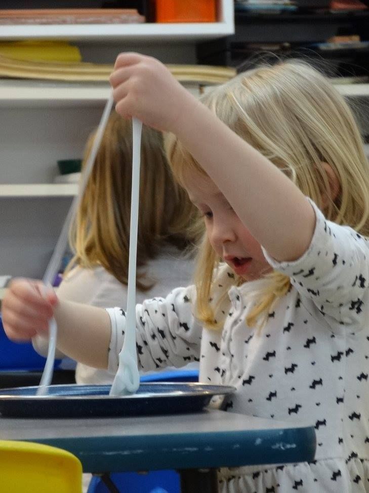 Girl with blonde hair pulling a long, white, slimy substance out of a tray at a table in a classroom