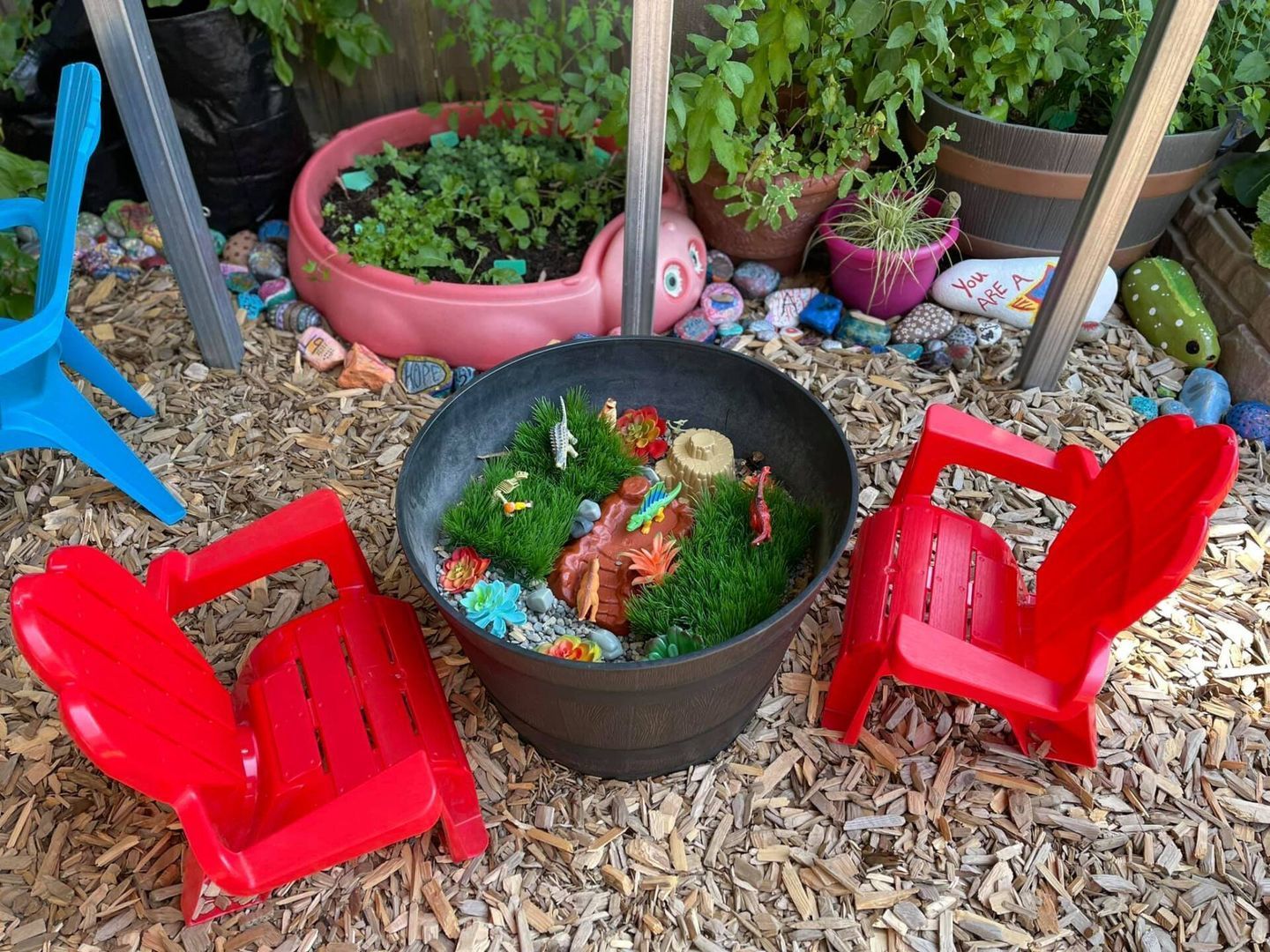 Child's outdoor play area with miniature garden in a pot, red chairs, and colorful decorations