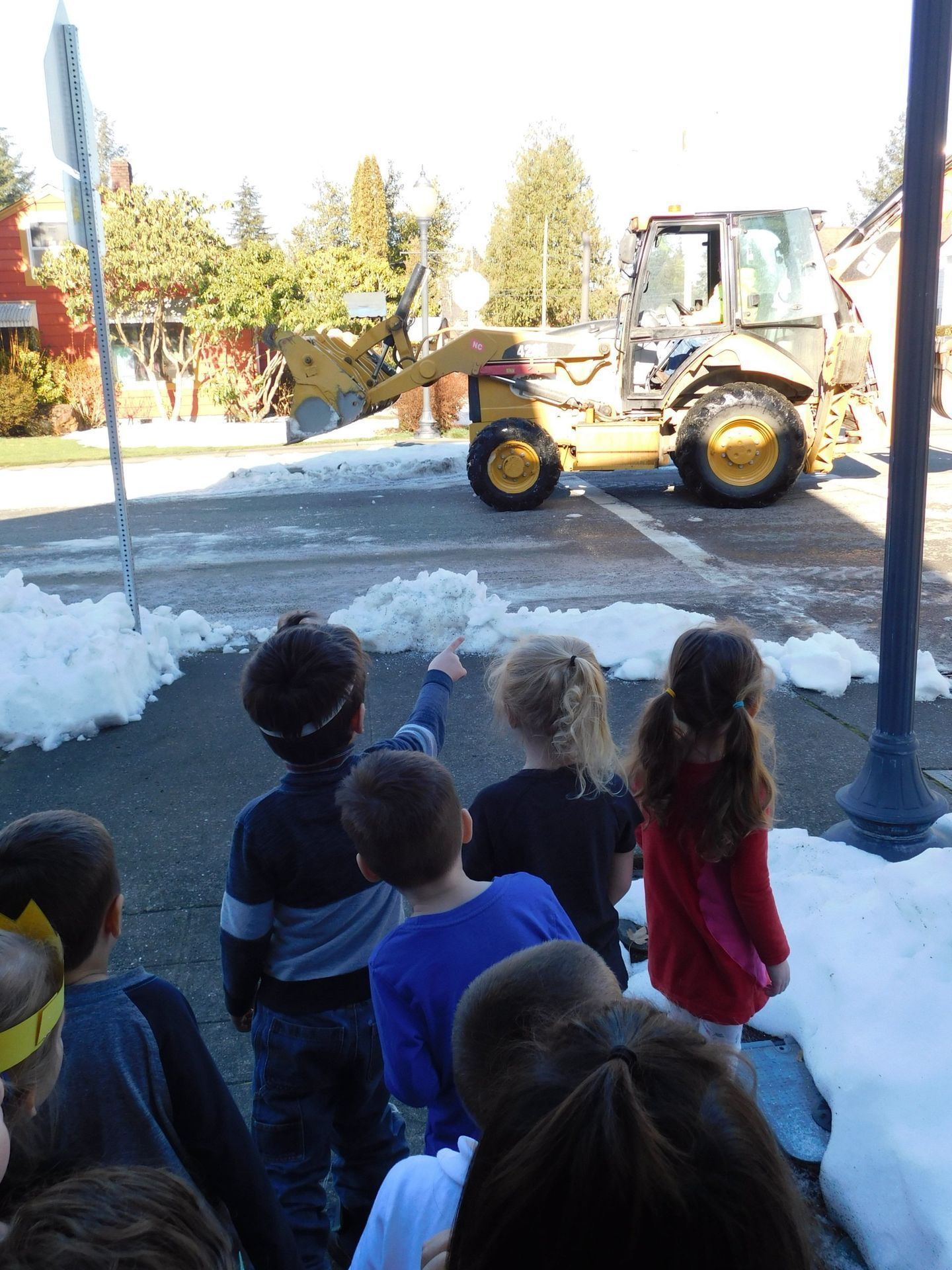 Children watch a yellow snowplow on a street clearing snow, pointing