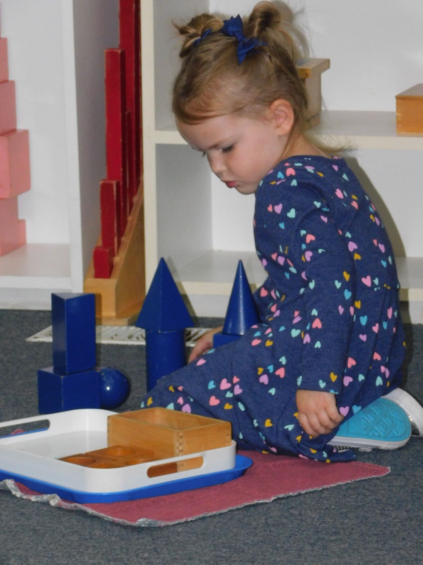 A young girl in blue dress playing with wooden geometric shapes on the floor