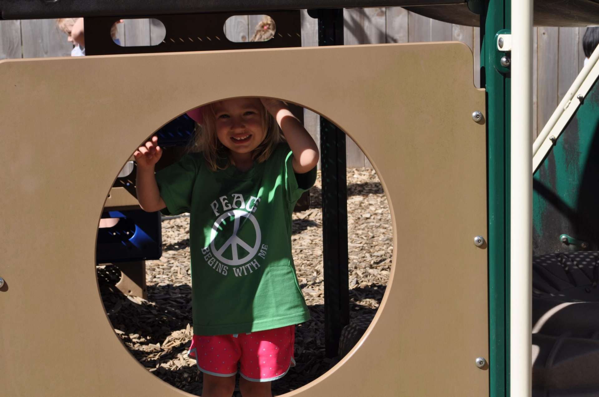 Young girl smiles through a circular opening in a playground wall, wearing green shirt and pink shorts